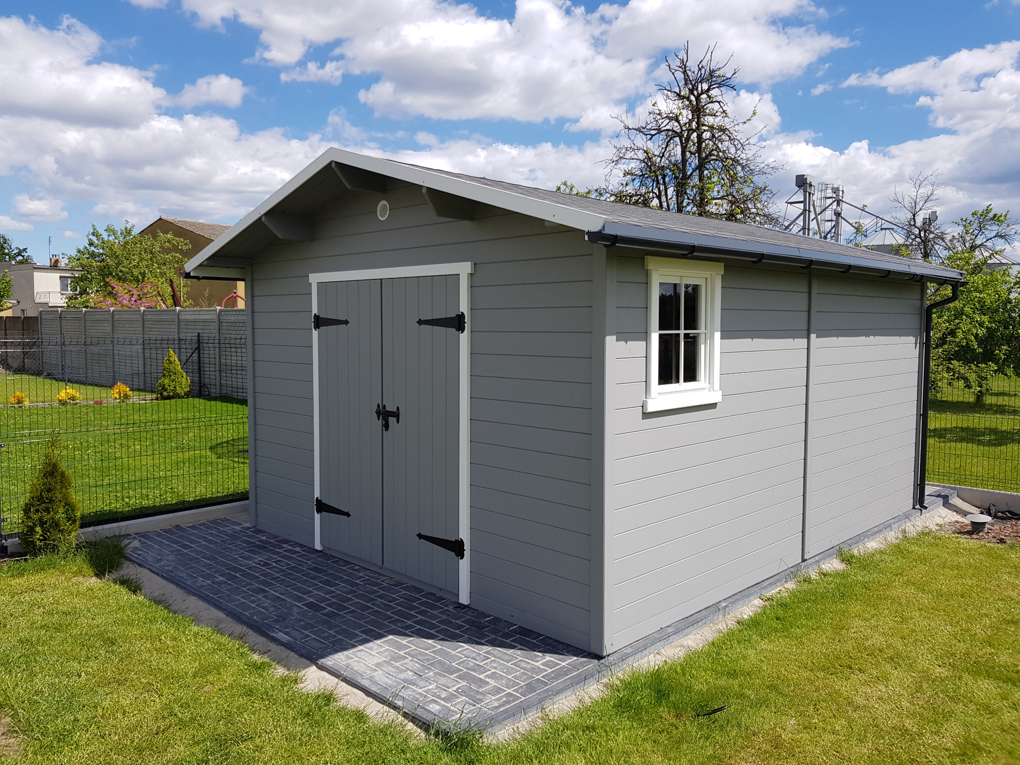 Szara, drewniana altana z białymi obramowaniami okien i czarnymi zawiasami,  umieszczona na kostce brukowej na tle zielonego trawnika i błękitnego nieba.