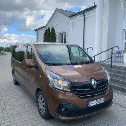 Brązowy bus Renault Trafic na tle białego budynku z kolumnami i schodami. Tablica rejestracyjna ESI 5900A. Pochmurne niebo w tle.