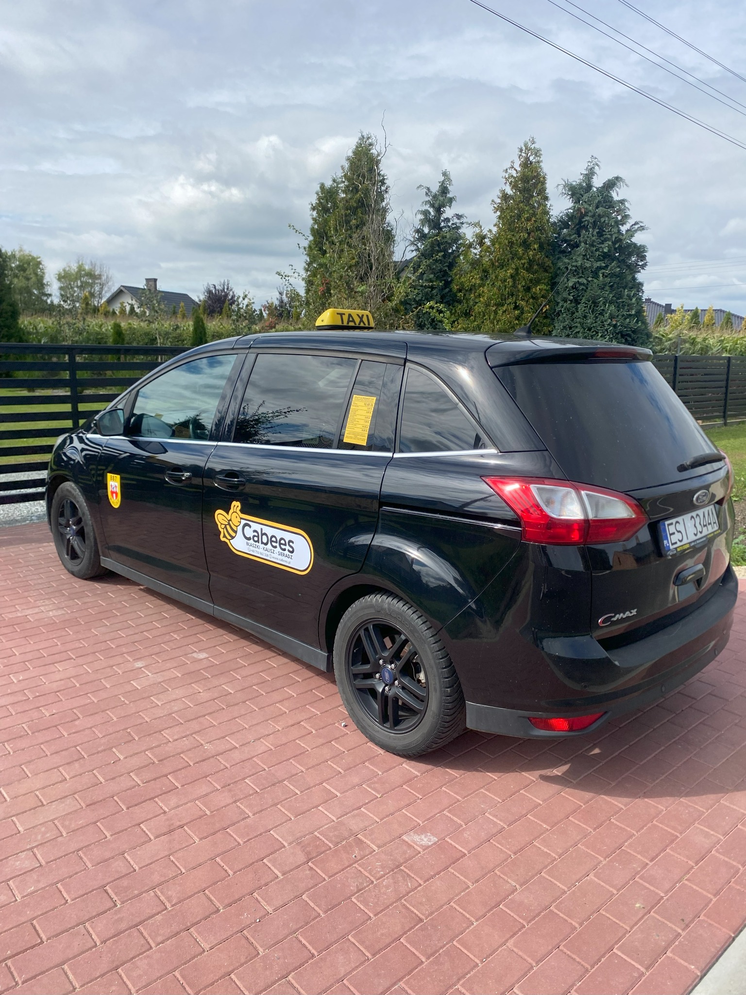 Czarny Ford C-Max Taxi 'Cabees' z żółtym szyldem na dachu, zaparkowany na czerwonej kostce brukowej. Widok z boku, na karoserii logo firmy i herb.
