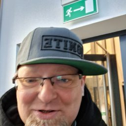 UT-R Marek Przyybylski - Mężczyzna w czapce z daszkiem i okularach, stojący pod zielonym znakiem ewakuacyjnym z piktogramem biegnącej osoby i strzałką w prawo.