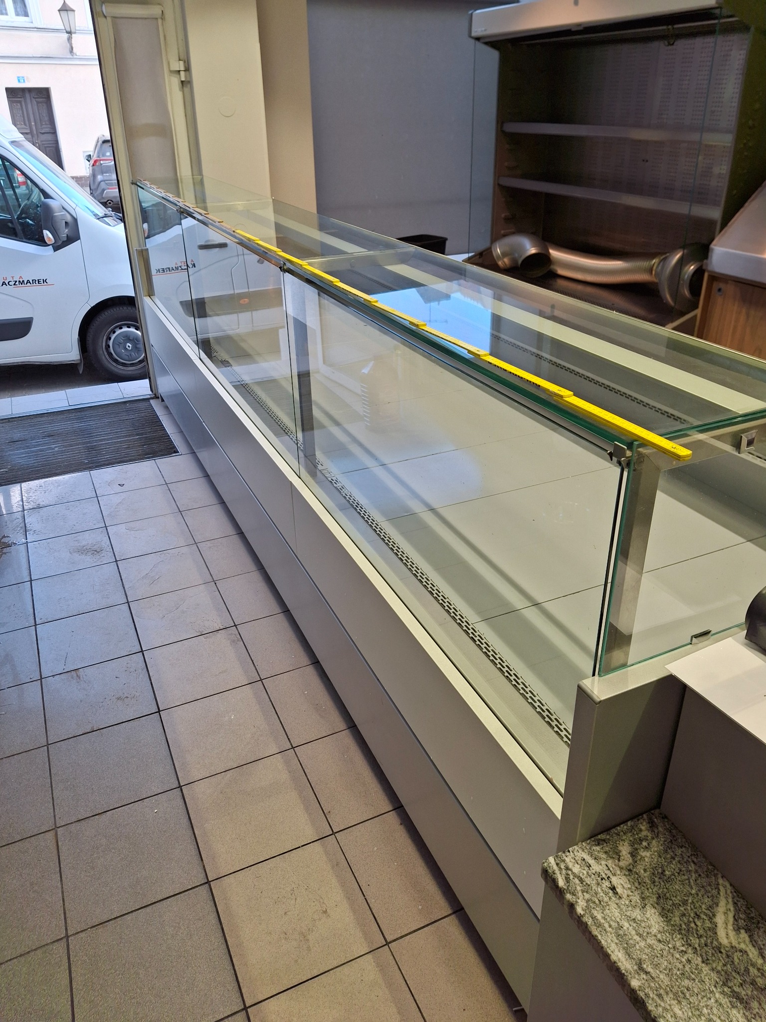 Szklana witryna chłodnicza z żółtymi elementami w lokalu, obok zaparkowany biały bus z logo firmy. Widoczna podłoga z płytek i fragment instalacji wentylacyjnej.