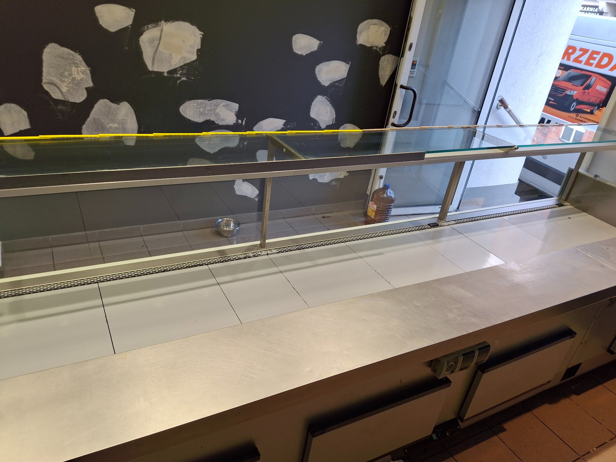 Półka chłodnicza ze stali nierdzewnej w lokalu, widoczne ślady po remoncie na ścianie i butelka z płynem. Widok na drzwi wejściowe z naklejką reklamową.