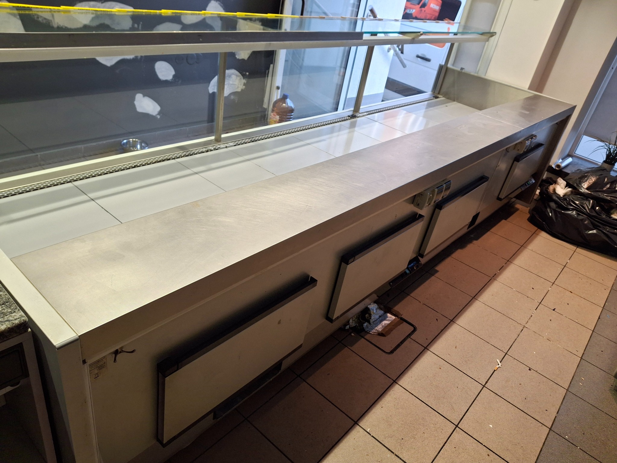 Chłodnicza lada ekspozycyjna ze stali nierdzewnej, widoczna z boku, z czarnymi workami na śmieci w tle, na brudnej podłodze z płytek.