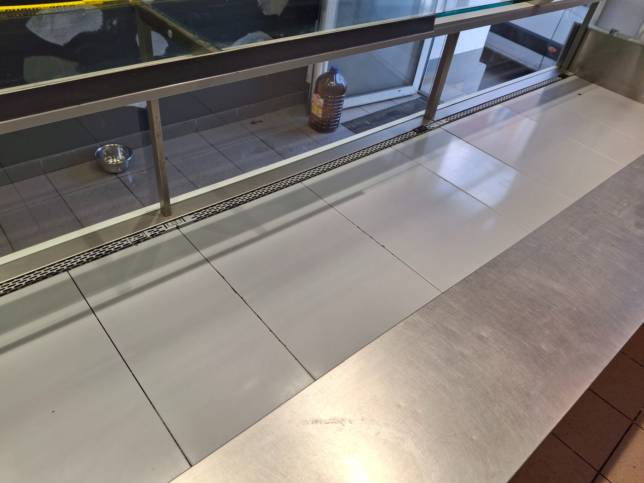 Półka w chłodni z metalową miską i butelką oleju w tle, widok z góry. Czysta, wyłożona płytkami podłoga. Minimalistyczny wystrój.