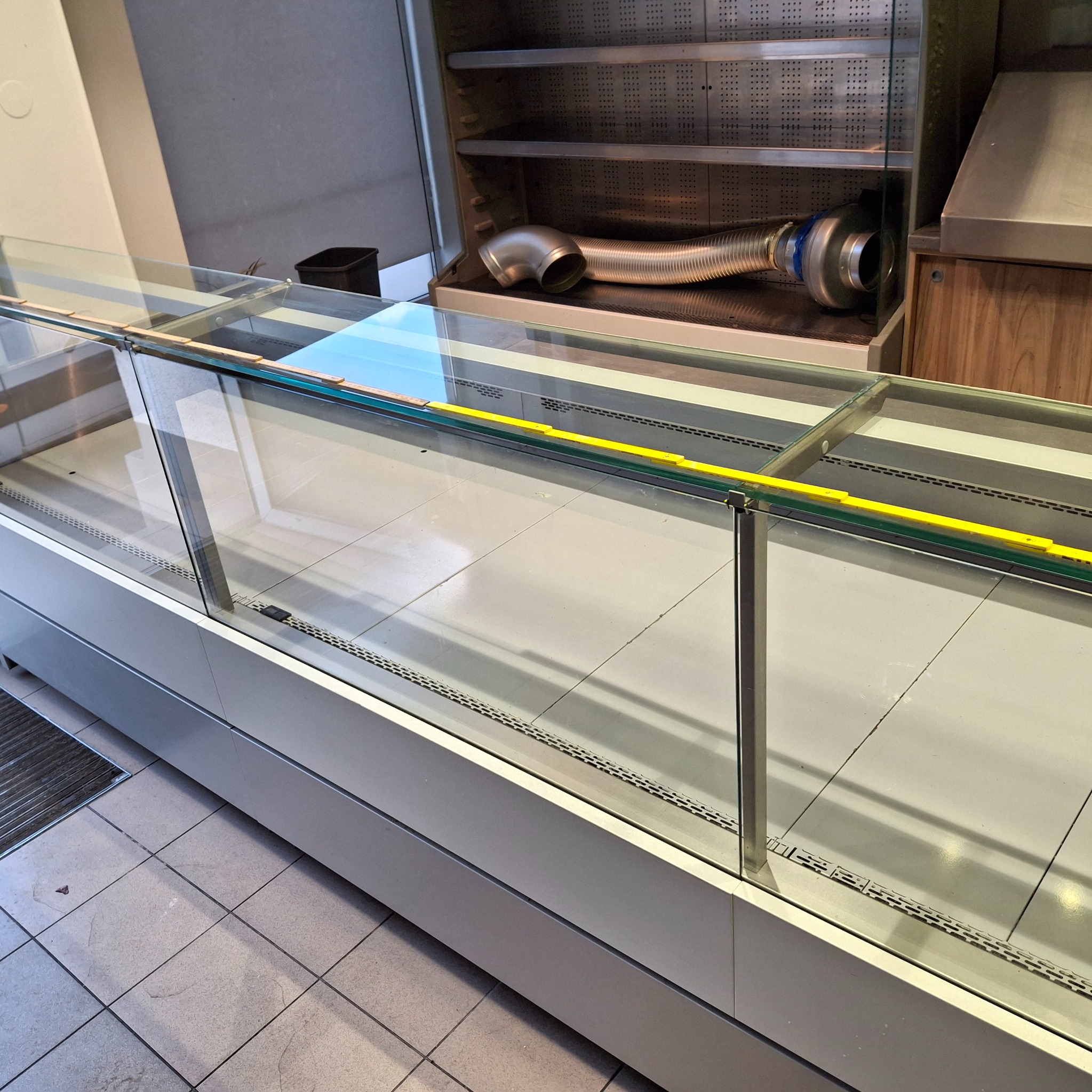 Pusta, szklana witryna chłodnicza po demontażu, z widocznym systemem wentylacji w tle. Posadzka z płytek. Widok z góry.