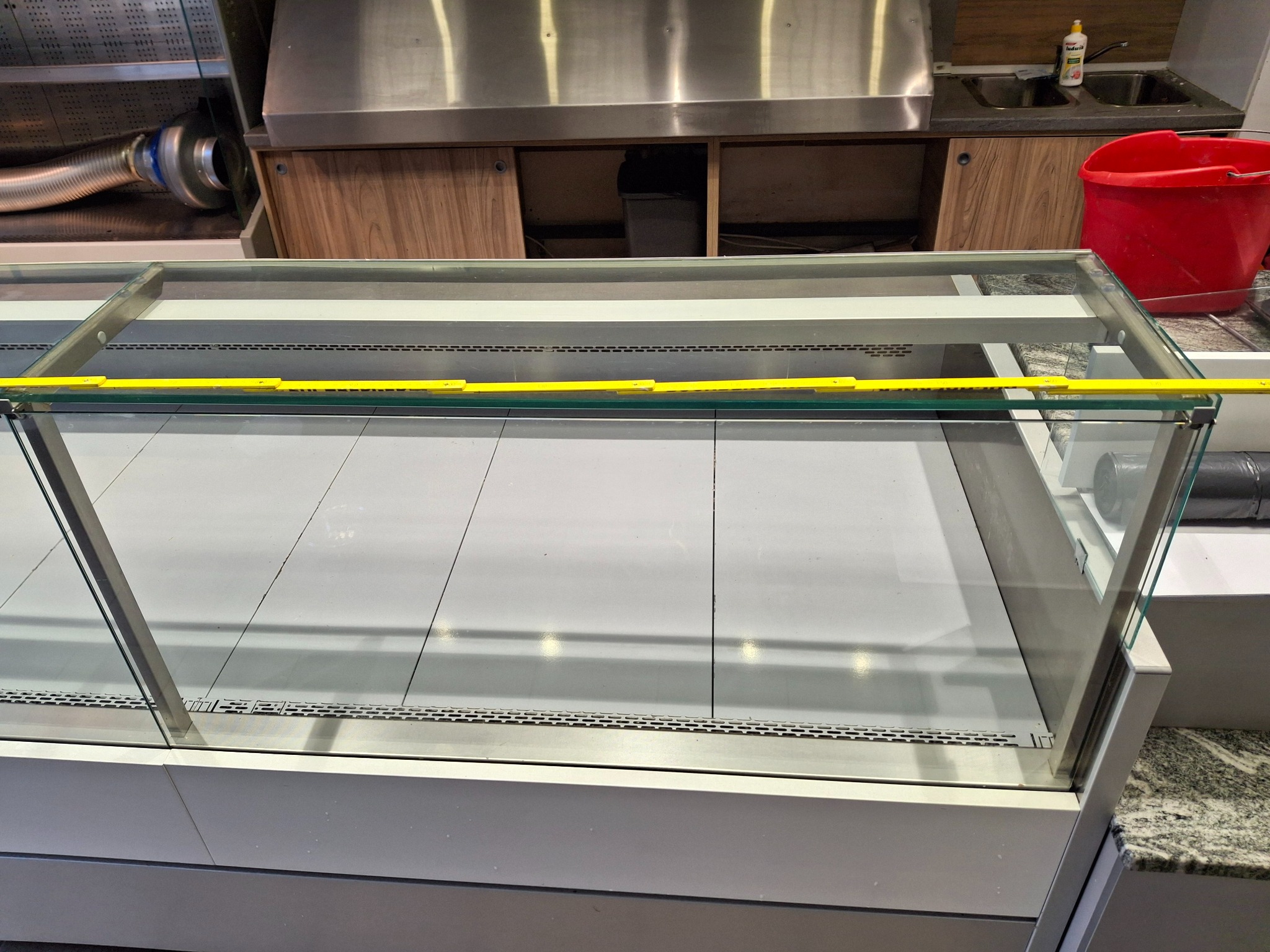 Pusta witryna chłodnicza z żółtą miarą, wnętrze lokalu gastronomicznego w tle. Widoczny fragment zlewu i czerwone wiadro, sugerujące przygotowania do sprzątania.