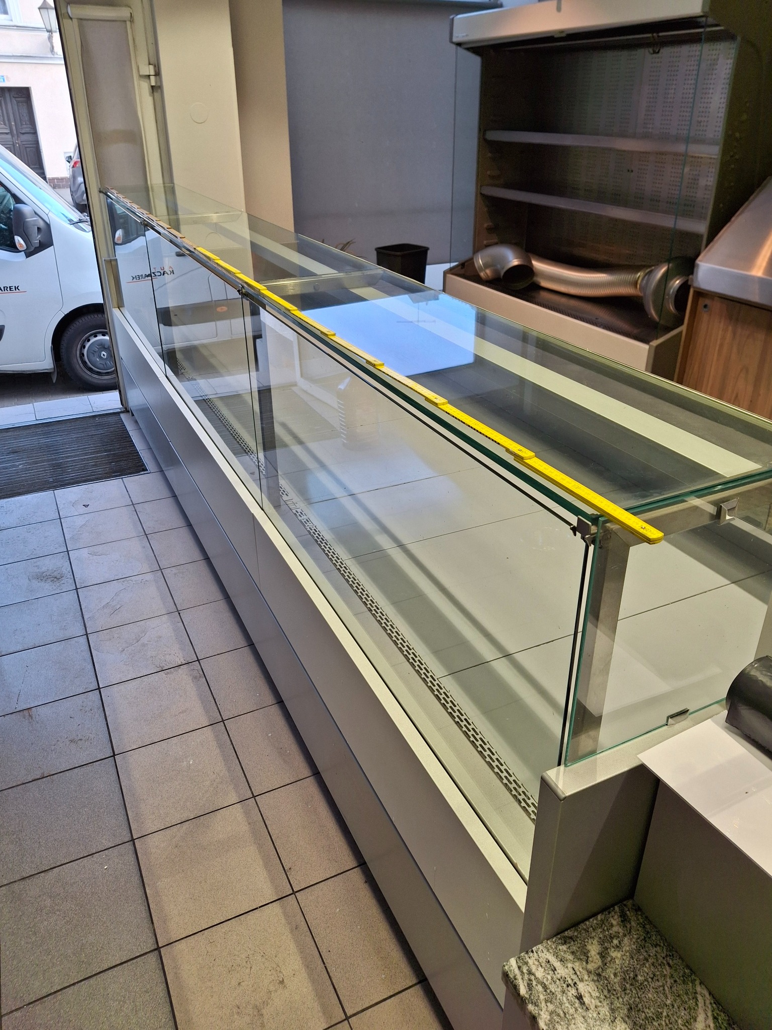 Pusta, szklana witryna chłodnicza z żółtą miarką na krawędzi, ustawiona w lokalu. Na zewnątrz widoczny bus dostawczy. Lokalizacja: wnętrze.