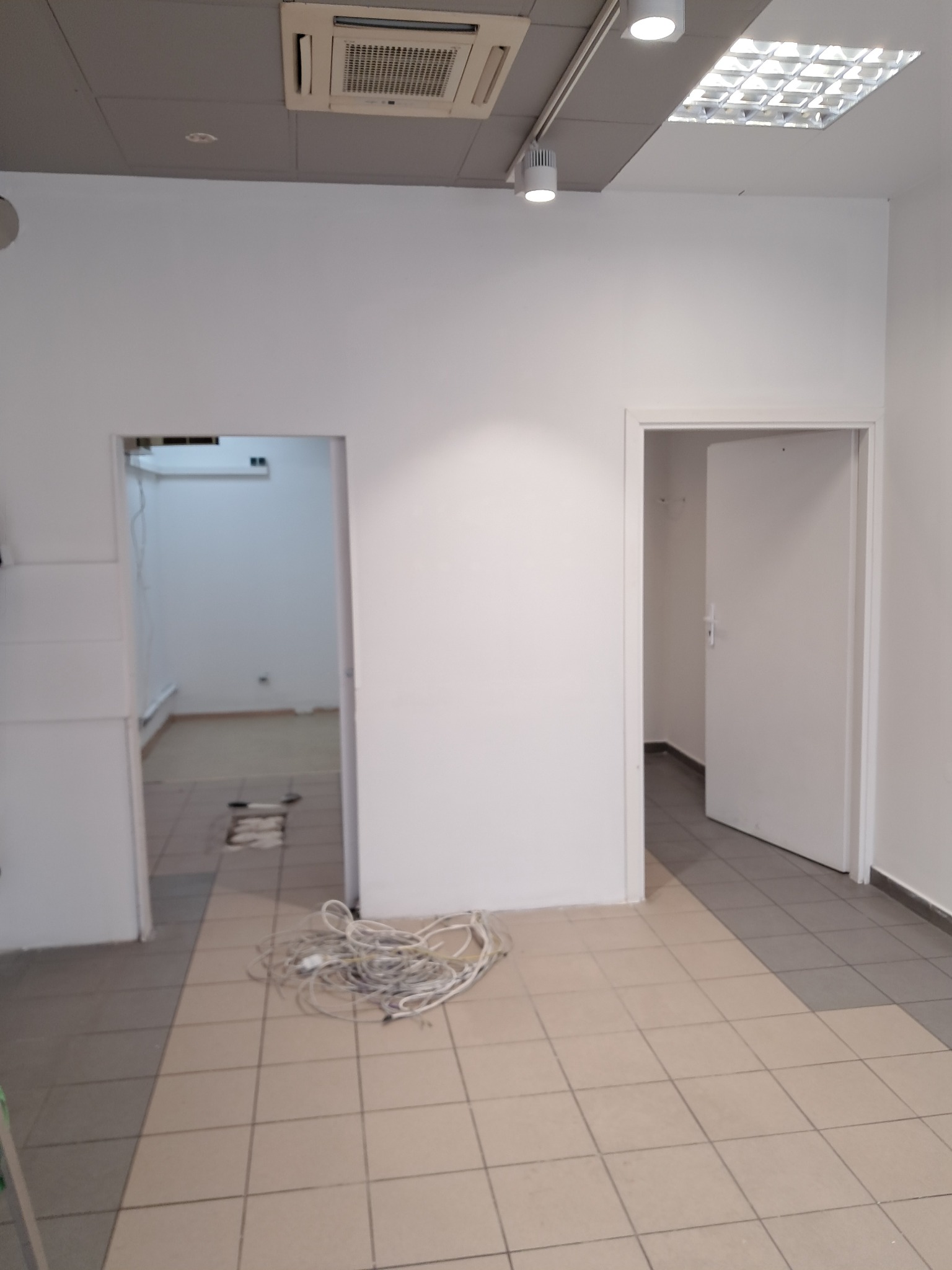 Puste, białe pomieszczenie z dwoma otwartymi drzwiami, widoczne kable na podłodze. Sufit z klimatyzacją i oświetleniem. Pomieszczenie po opróżnianiu.