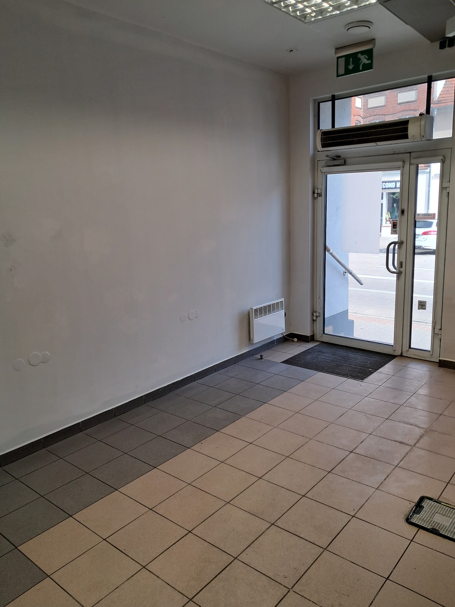 Pusta przestrzeń biurowa z białymi ścianami, jasną podłogą z płytek i szklanymi drzwiami wejściowymi. Widoczny grzejnik na ścianie i znak ewakuacyjny.