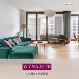 Jasne wnętrze mieszkania z zieloną sofą narożną, stolikiem kawowym i wyjściem na balkon. Widoczne okna balkonowe z drewnianymi ramami i budynek w tle.