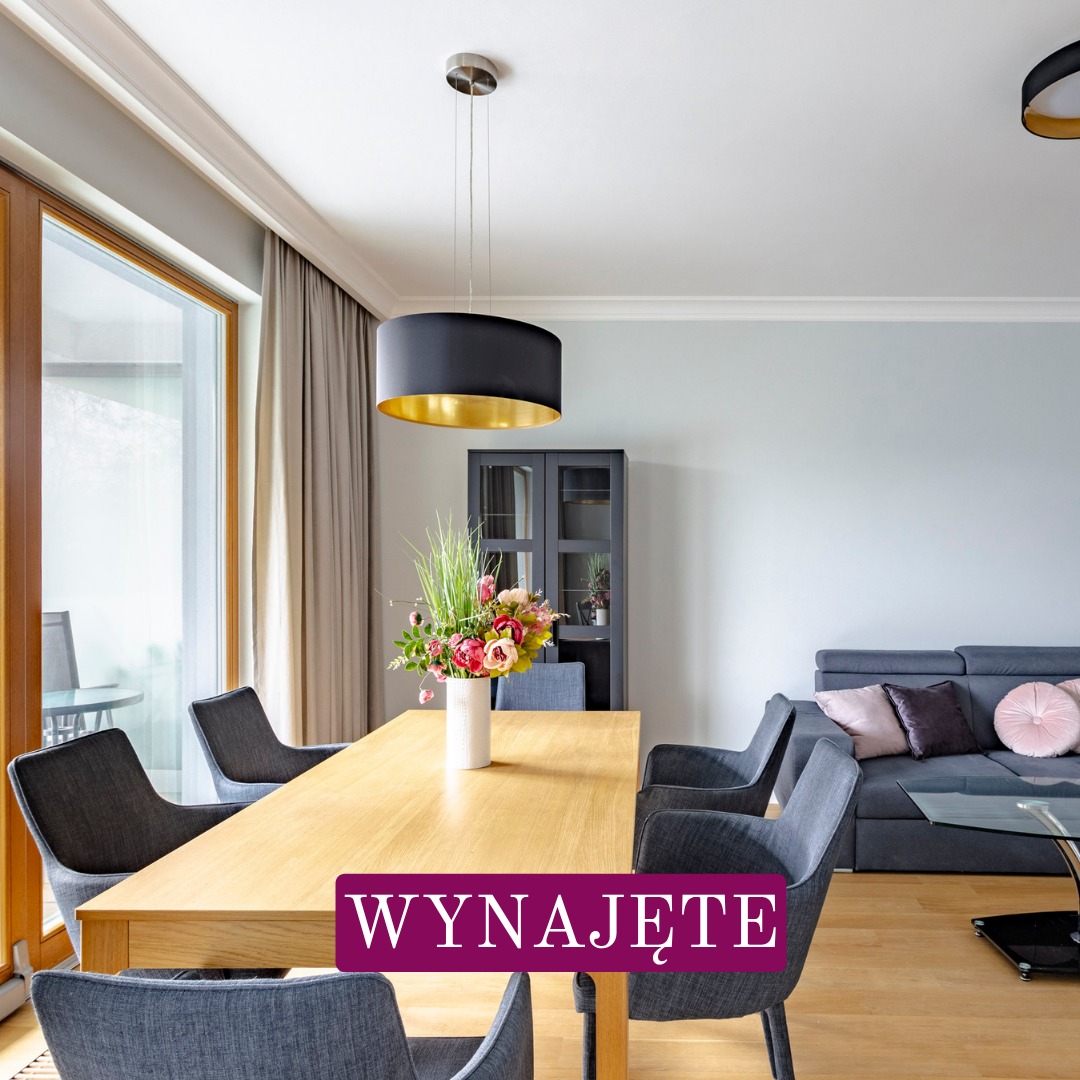 Jasny salon z drewnianym stołem, krzesłami, szarą sofą i kwiatami. Nad stołem wisi czarna lampa z złotym wnętrzem. Na zdjęciu widnieje napis 'WYNAJĘTE'.