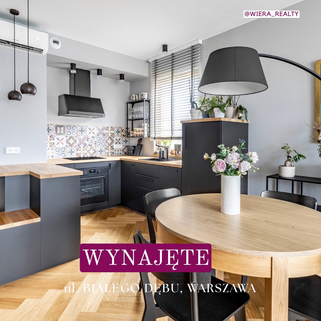 Nowoczesna kuchnia w szarościach z drewnianym blatem i parkietem w jodełkę. Na stole wazon z kwiatami. Okno z żaluzjami. Napis 'Wynajęte'.