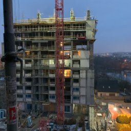 Monolit Sp. z o. o. - Wysoki budynek żelbetowy w trakcie budowy, widoczny czerwony dźwig i pracownicy na górnych kondygnacjach. W tle zabudowa miejska i rzeka, wieczorny krajobraz.