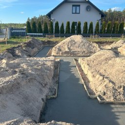 Monolit Sp. z o. o. - Wylane fundamenty pod dom jednorodzinny, widoczne szalunki i świeży beton. W tle dom w stylu nowoczesnej stodoły i rząd tuj przy ogrodzeniu.