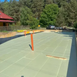 Fundamenty domu z izolacją termiczną, przygotowane do dalszych prac. Widoczne pionowe rury instalacyjne i obrzeża szalunkowe. W tle las i fragment budynku.