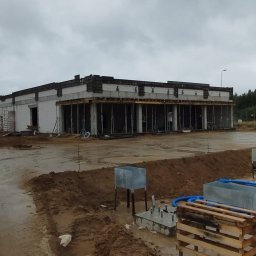 Monolit Sp. z o. o. - Budowa hali w trakcie realizacji, widoczne fundamenty, ściany z białego bloczka i podpory szalunkowe. W tle pochmurne niebo i drzewa.