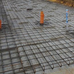 Monolit Sp. z o. o. - Zbrojenie płyty fundamentowej z wystającymi rurami kanalizacyjnymi. Widoczne siatki zbrojeniowe i betonowe podkładki dystansowe. Prace budowlane w toku.