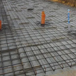 Zbrojenie płyty fundamentowej z wystającymi rurami kanalizacyjnymi. Widoczne siatki zbrojeniowe i betonowe podkładki dystansowe. Prace budowlane w toku.