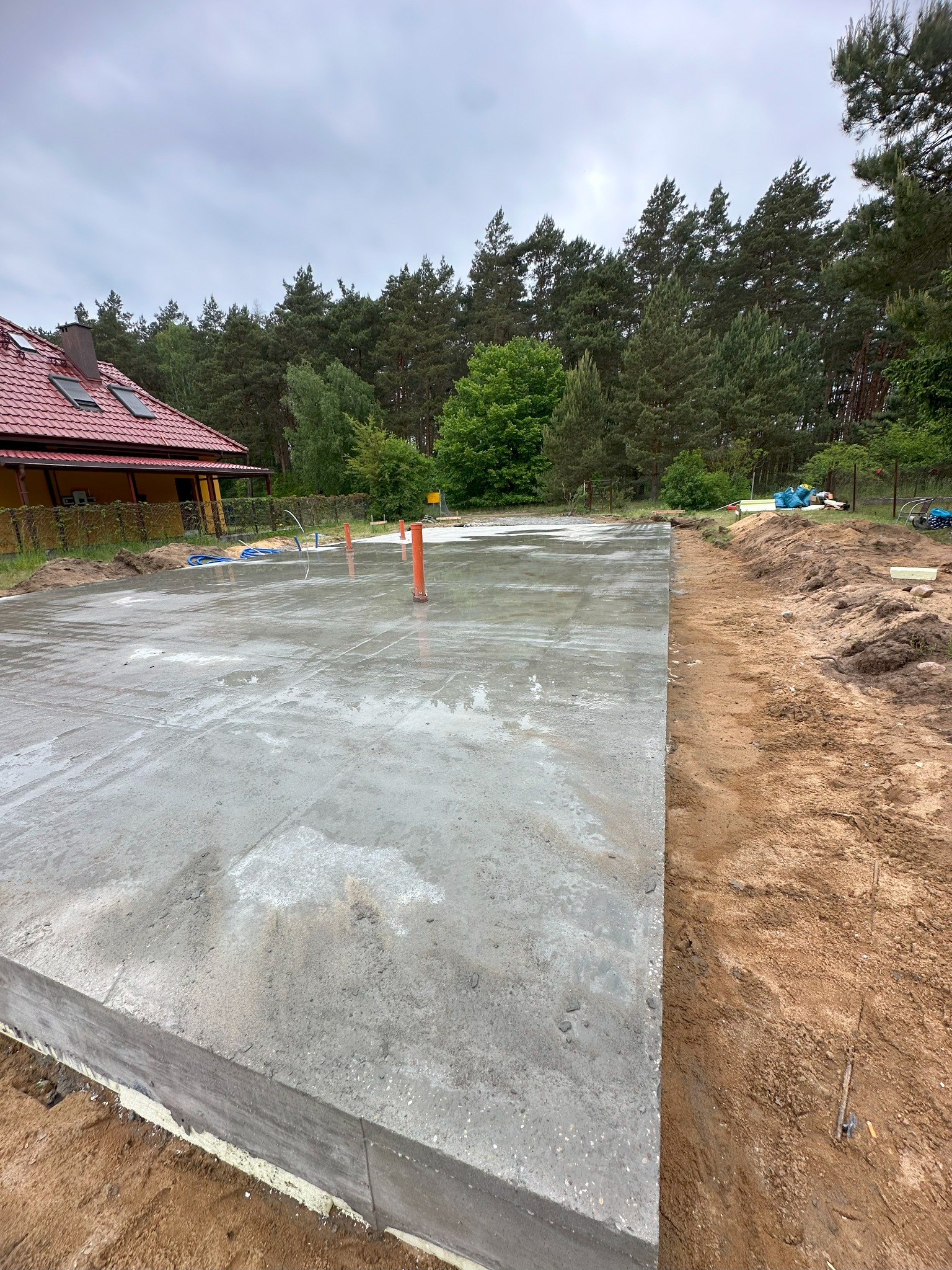 Świeżo wylana płyta fundamentowa w Bydgoszczy, widoczne pionowe rury instalacyjne, izolacja obwodowa, w tle dom z czerwoną dachówką i drzewa.