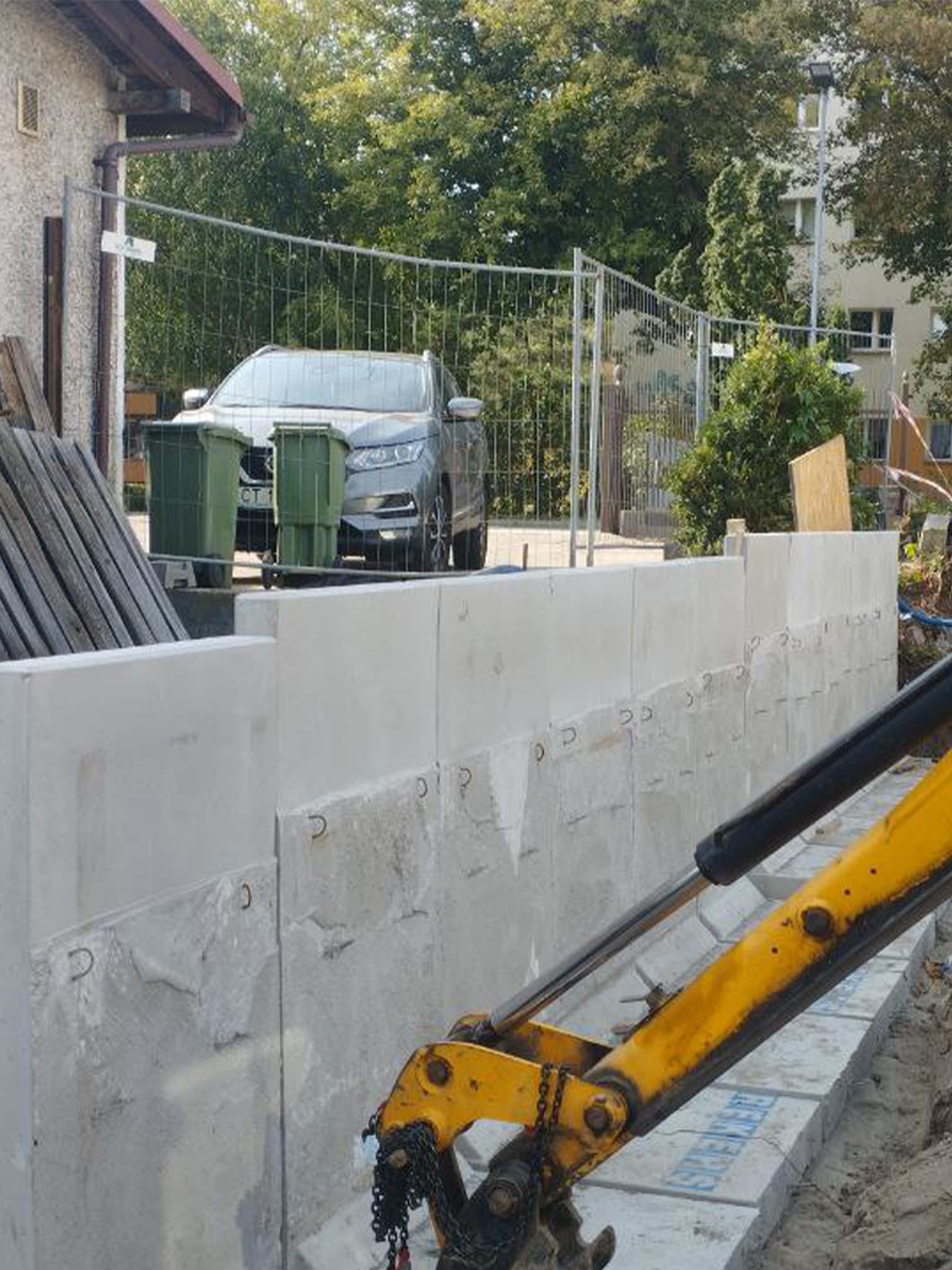 Wznoszenie muru oporowego z prefabrykatów betonowych na posesji, widoczne elementy koparki i ogrodzenie. W tle zaparkowany samochód osobowy.