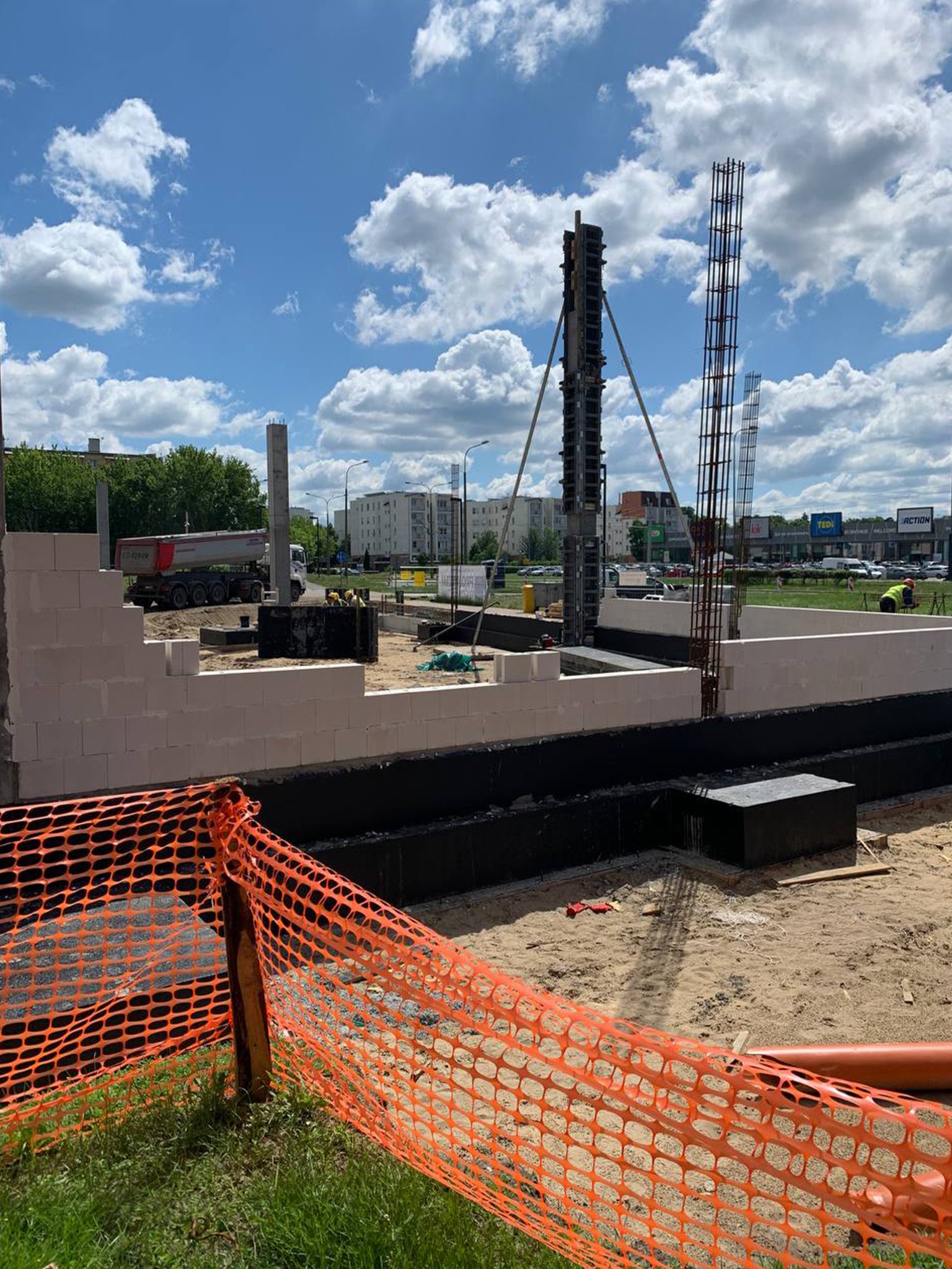 Budowa domu: fundamenty z izolacją, częściowo wymurowane ściany z betonu komórkowego, zbrojenie kolumn. Na drugim planie ciężarówka i budynki mieszkalne.