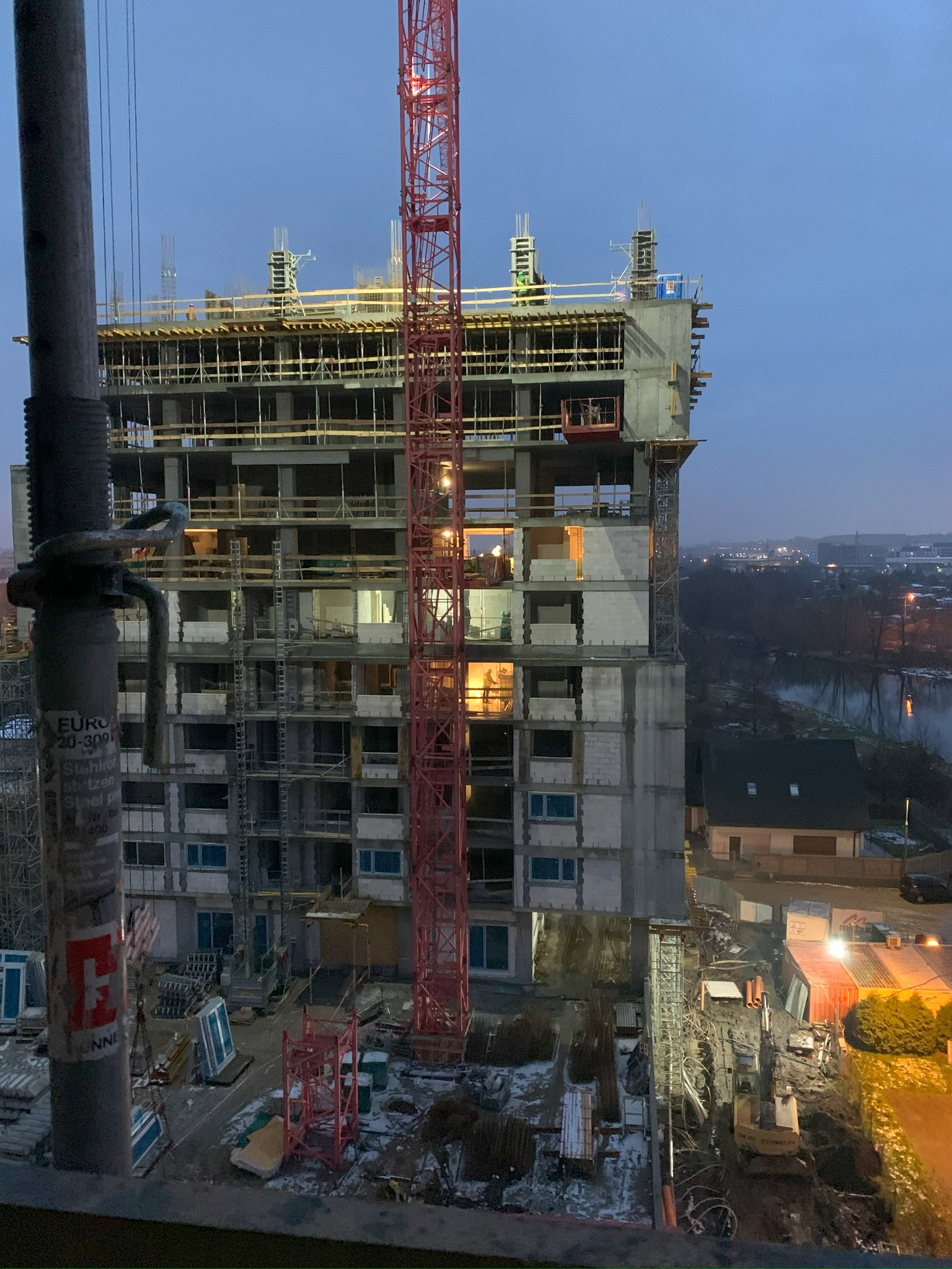 Wysoki budynek żelbetowy w trakcie budowy, widoczny czerwony dźwig i pracownicy na górnych kondygnacjach. W tle zabudowa miejska i rzeka, wieczorny krajobraz.