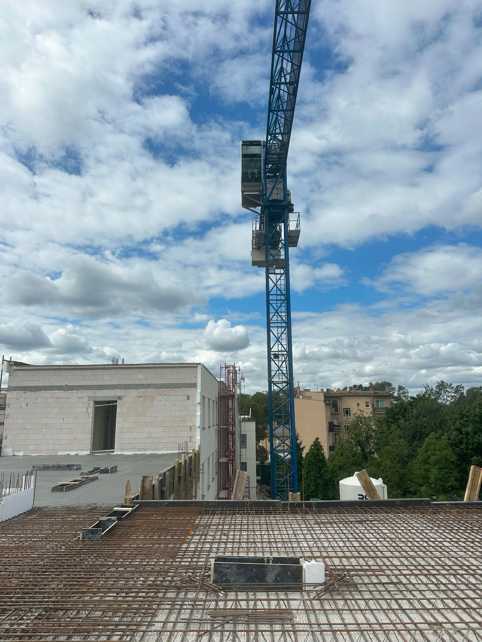 Zbrojenie stropu budynku w trakcie budowy, widok z góry na siatkę zbrojeniową, w tle budynek w stanie surowym otwartym i żuraw budowlany.