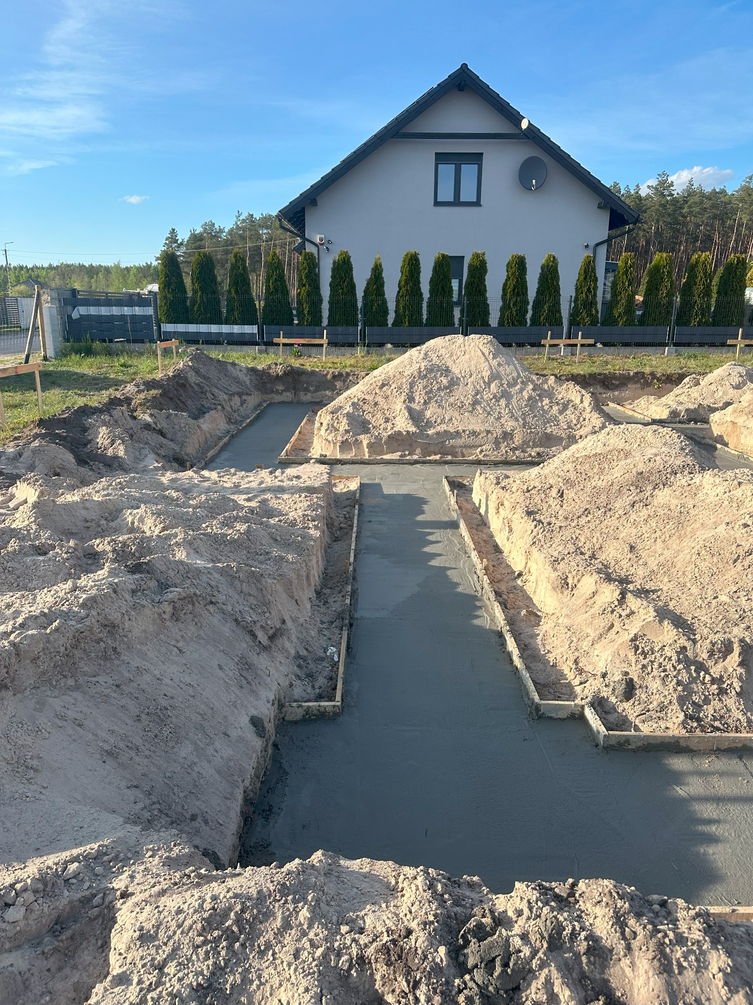 Wylane fundamenty pod dom jednorodzinny, widoczne szalunki i świeży beton. W tle dom w stylu nowoczesnej stodoły i rząd tuj przy ogrodzeniu.