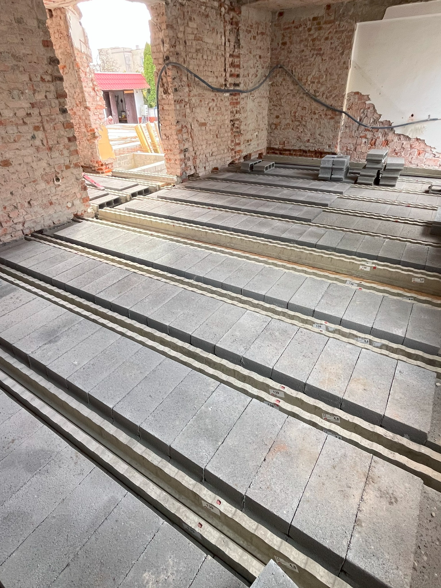 Ułożone bloczki stropowe na legarach w surowym wnętrzu budynku z ceglanymi ścianami. Widoczne instalacje. Fragment budynku z czerwonym dachem w tle.