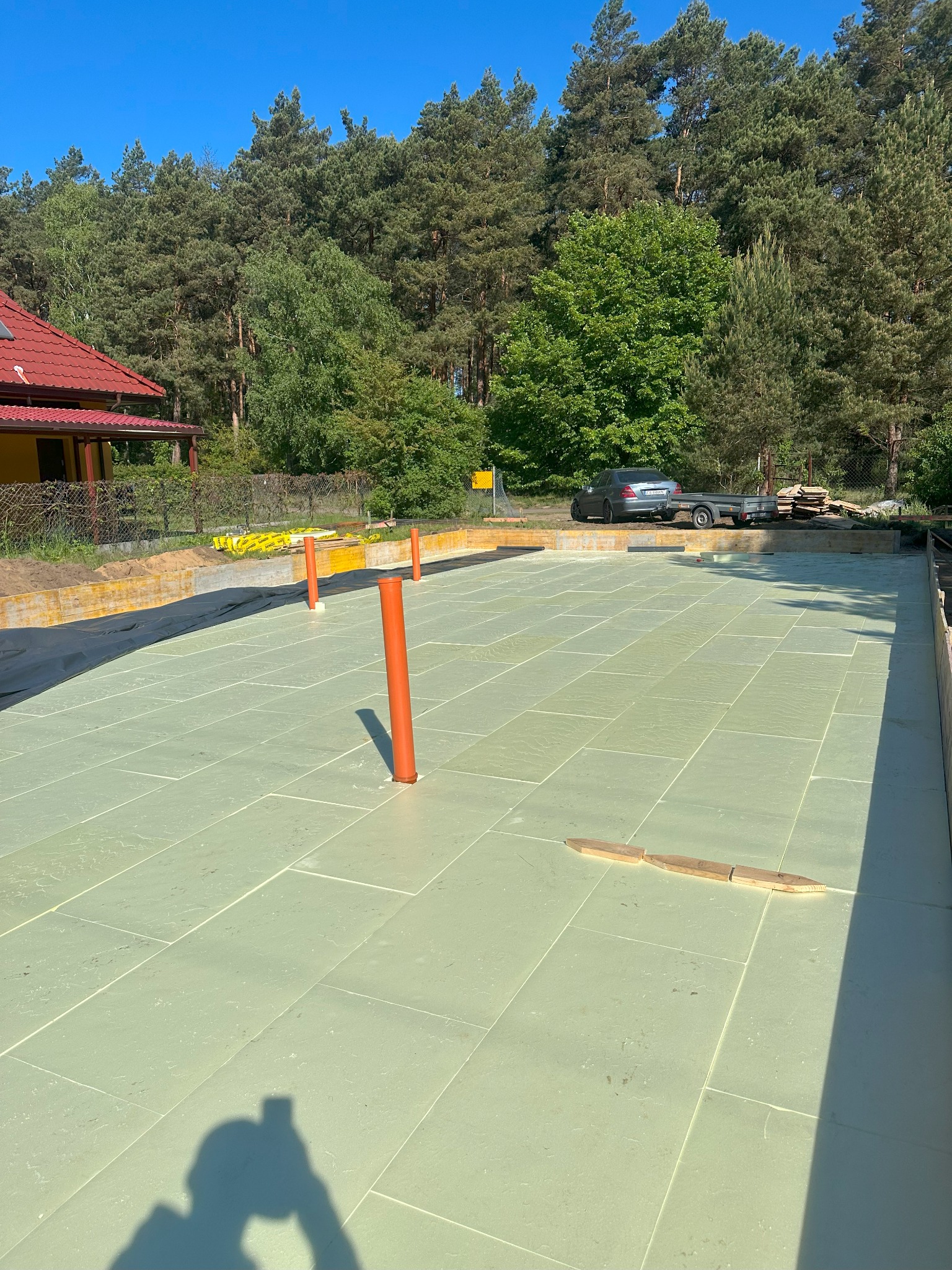 Fundamenty domu z izolacją termiczną, przygotowane do dalszych prac. Widoczne pionowe rury instalacyjne i obrzeża szalunkowe. W tle las i fragment budynku.