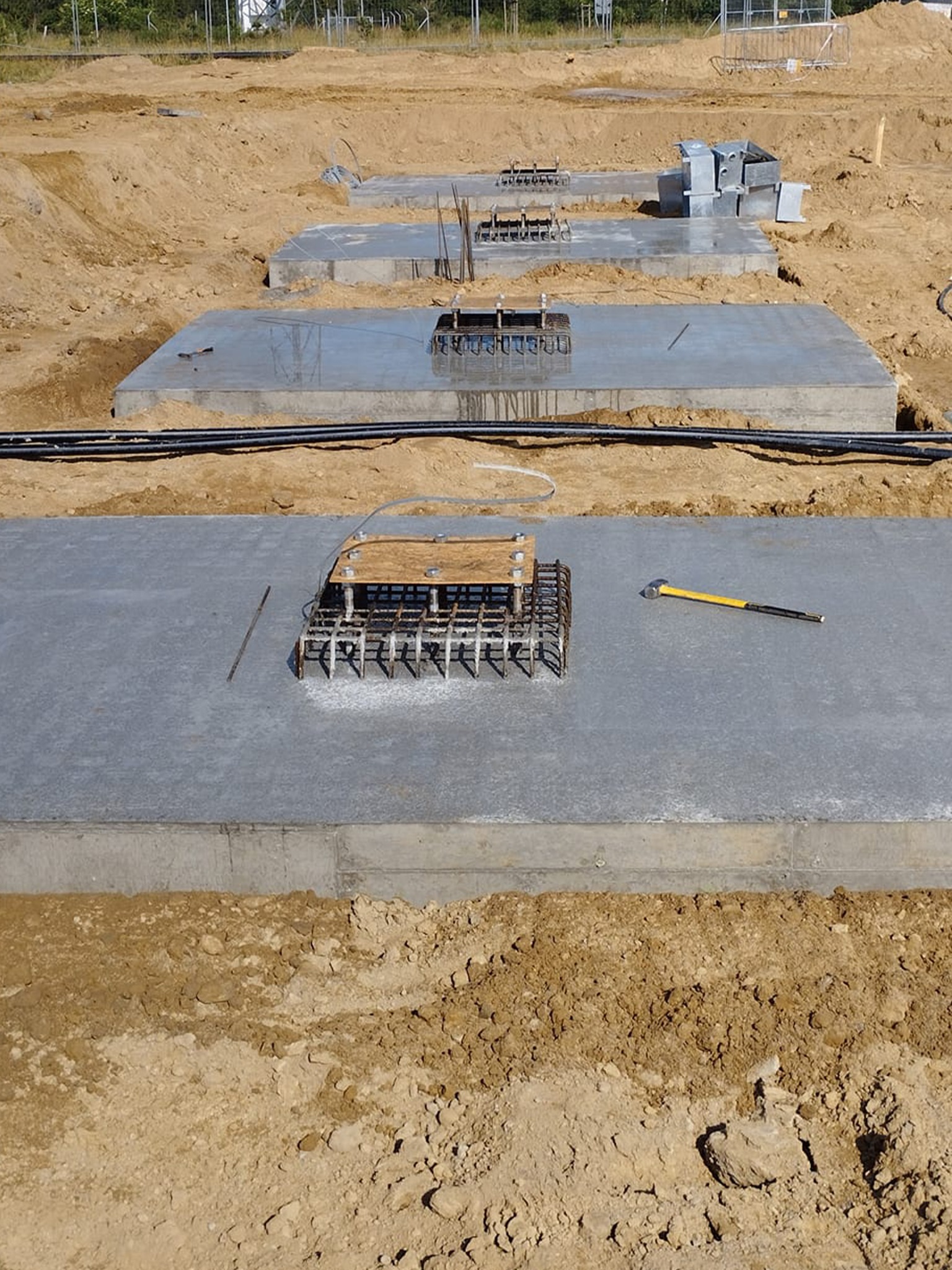 Betonowe fundamenty z wystającą zbrojeniem, widoczne czarne rury i młotek na jednym z fundamentów, teren budowy w tle, ziemia, wczesny etap budowy.