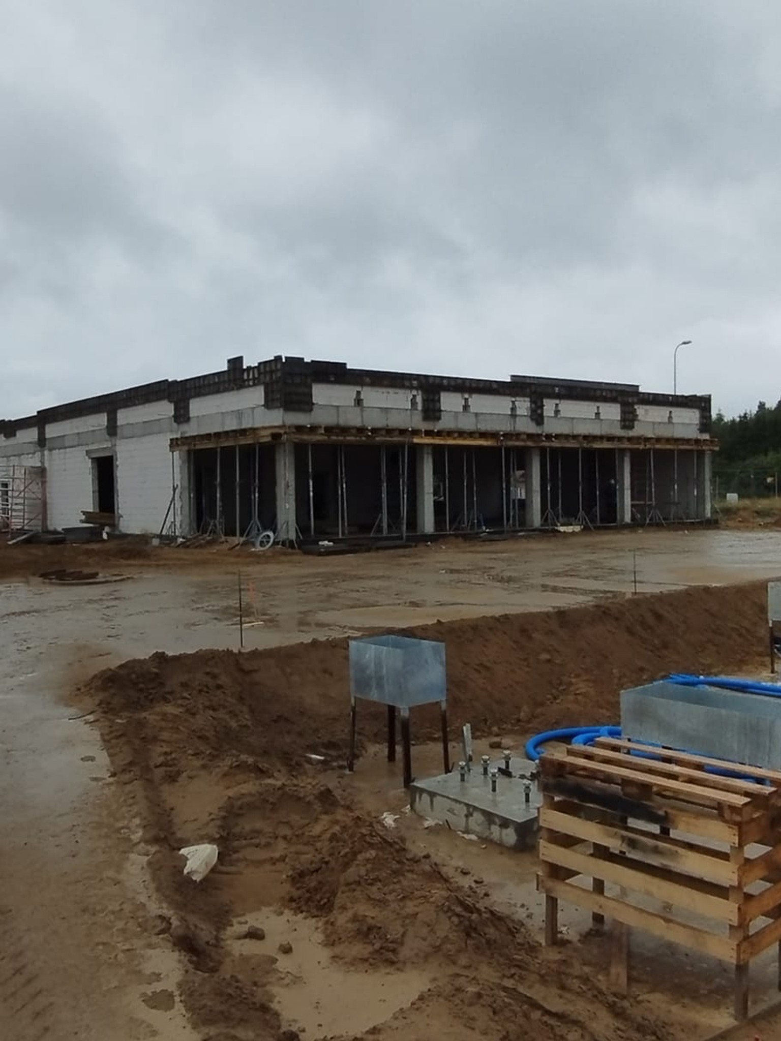 Budowa hali w trakcie realizacji, widoczne fundamenty, ściany z białego bloczka i podpory szalunkowe. W tle pochmurne niebo i drzewa.