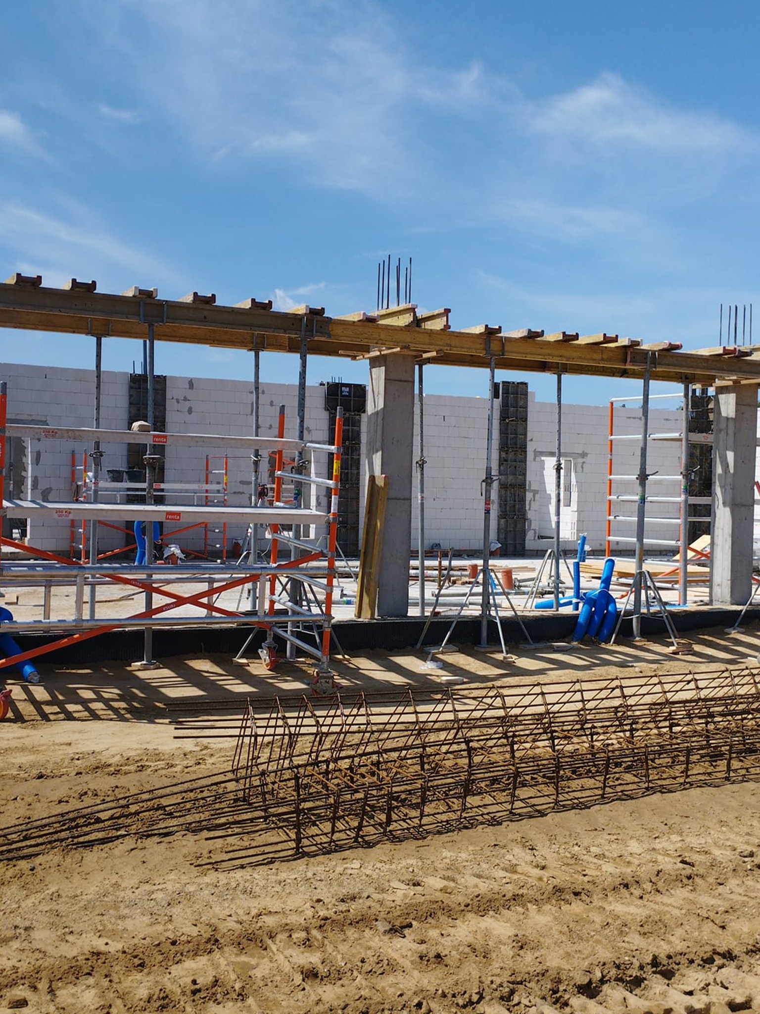 Budowa domu z betonu komórkowego: zbrojenie fundamentów, szalunki, rusztowania na tle błękitnego nieba. Widoczne elementy konstrukcyjne i postęp prac.