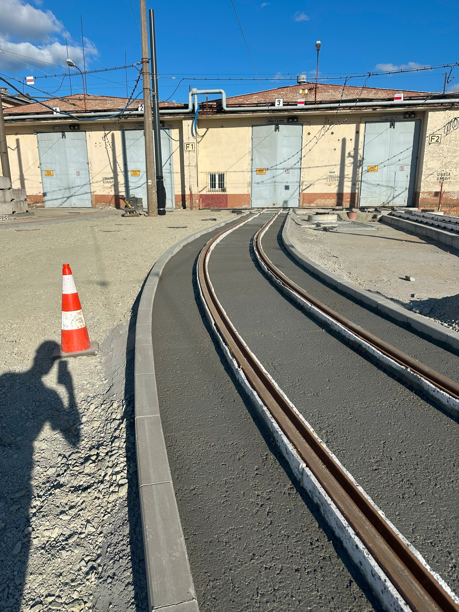 Nowo wyremontowane tory tramwajowe z betonowym wypełnieniem między szynami, pomarańczowy pachołek i cień osoby w rogu kadru, budynek z numerami nad bramami.
