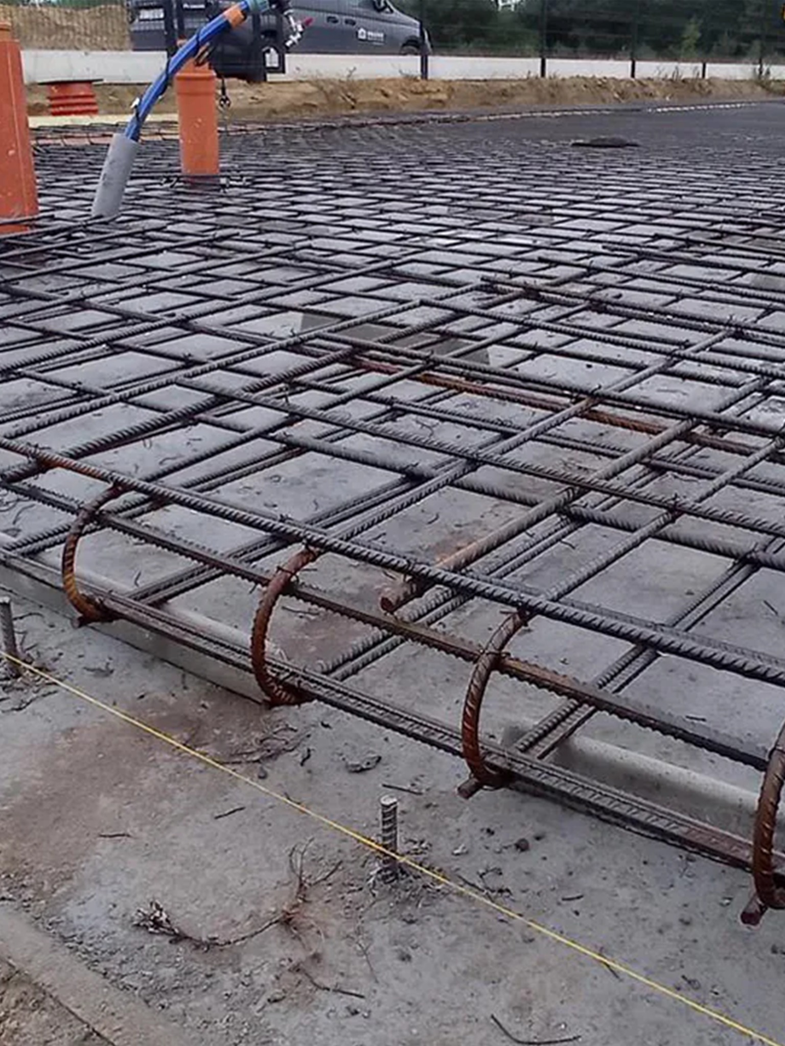 Zbrojenie płyty fundamentowej z widocznymi rurami instalacyjnymi i żółtą linką wyznaczającą poziom, na betonowej powierzchni. W tle widoczny samochód dostawczy.