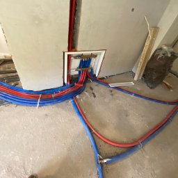 MIK-INSTAL Norbert Dembiński - Rozłożone, czerwono-niebieskie rury instalacji ogrzewania podłogowego, prowadzące do rozdzielacza w ścianie. Widoczne mocowania i fragment buta w dolnym rogu.