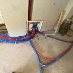 Rozłożone, czerwono-niebieskie rury instalacji ogrzewania podłogowego, prowadzące do rozdzielacza w ścianie. Widoczne mocowania i fragment buta w dolnym rogu.