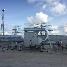 MIK-INSTAL Norbert Dembiński - Instalacja wentylacyjna na dachu budynku z widoczną wieżą telekomunikacyjną i liniami wysokiego napięcia w tle. Mężczyzna siedzi na rurze wentylacyjnej.