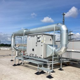 MIK-INSTAL Norbert Dembiński - Instalacja wentylacyjna na dachu budynku, z widocznymi przewodami wentylacyjnymi, wspornikami i elementami mocującymi. W tle błękitne niebo z chmurami.