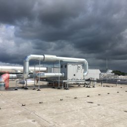 MIK-INSTAL Norbert Dembiński - Instalacja przemysłowa z srebrnymi rurami i szarą centralą wentylacyjną na płaskim dachu, widok z góry, pochmurne niebo w tle.