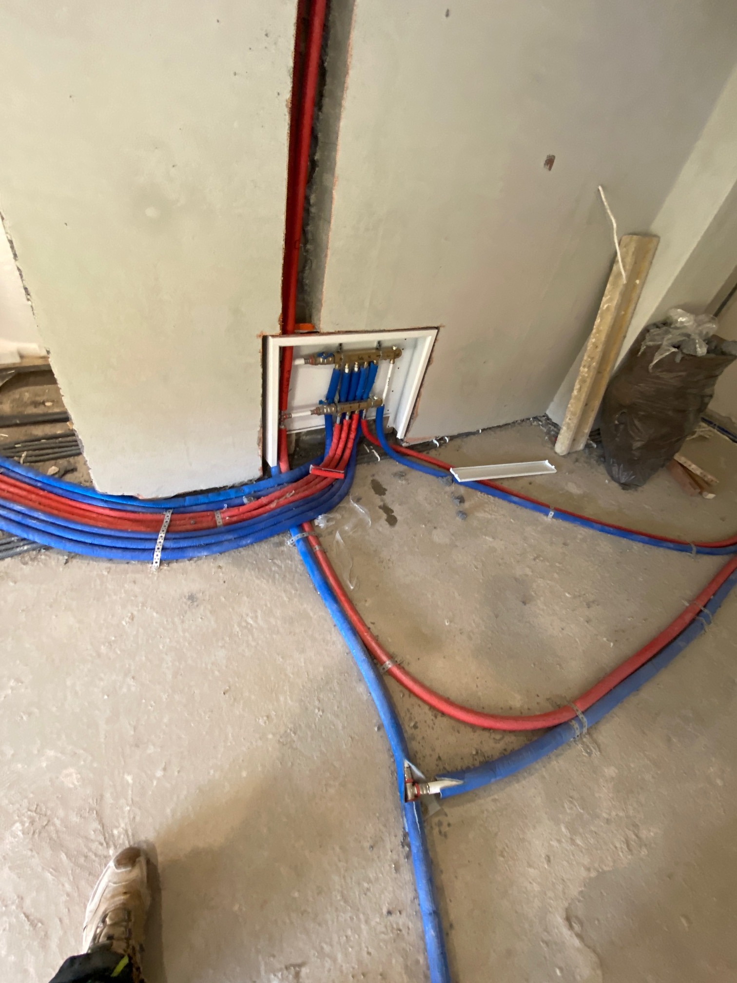 Rozłożone, czerwono-niebieskie rury instalacji ogrzewania podłogowego, prowadzące do rozdzielacza w ścianie. Widoczne mocowania i fragment buta w dolnym rogu.