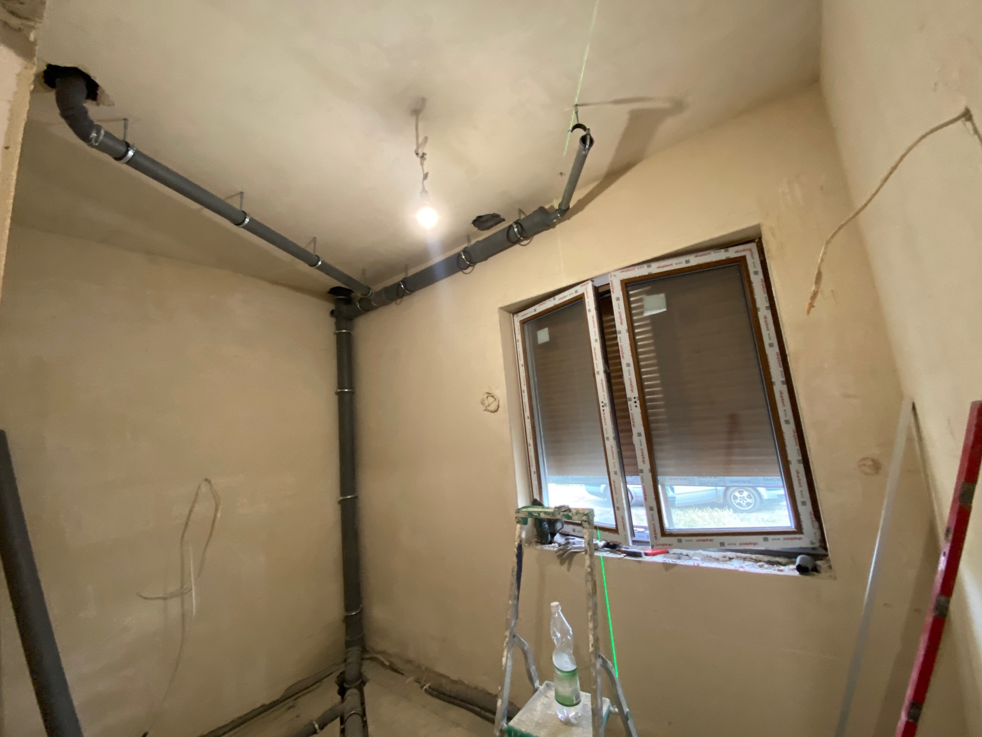 Szare rury kanalizacyjne zamontowane pod sufitem w surowym wnętrzu łazienki, widoczne narzędzia i drabina, okno z roletami.