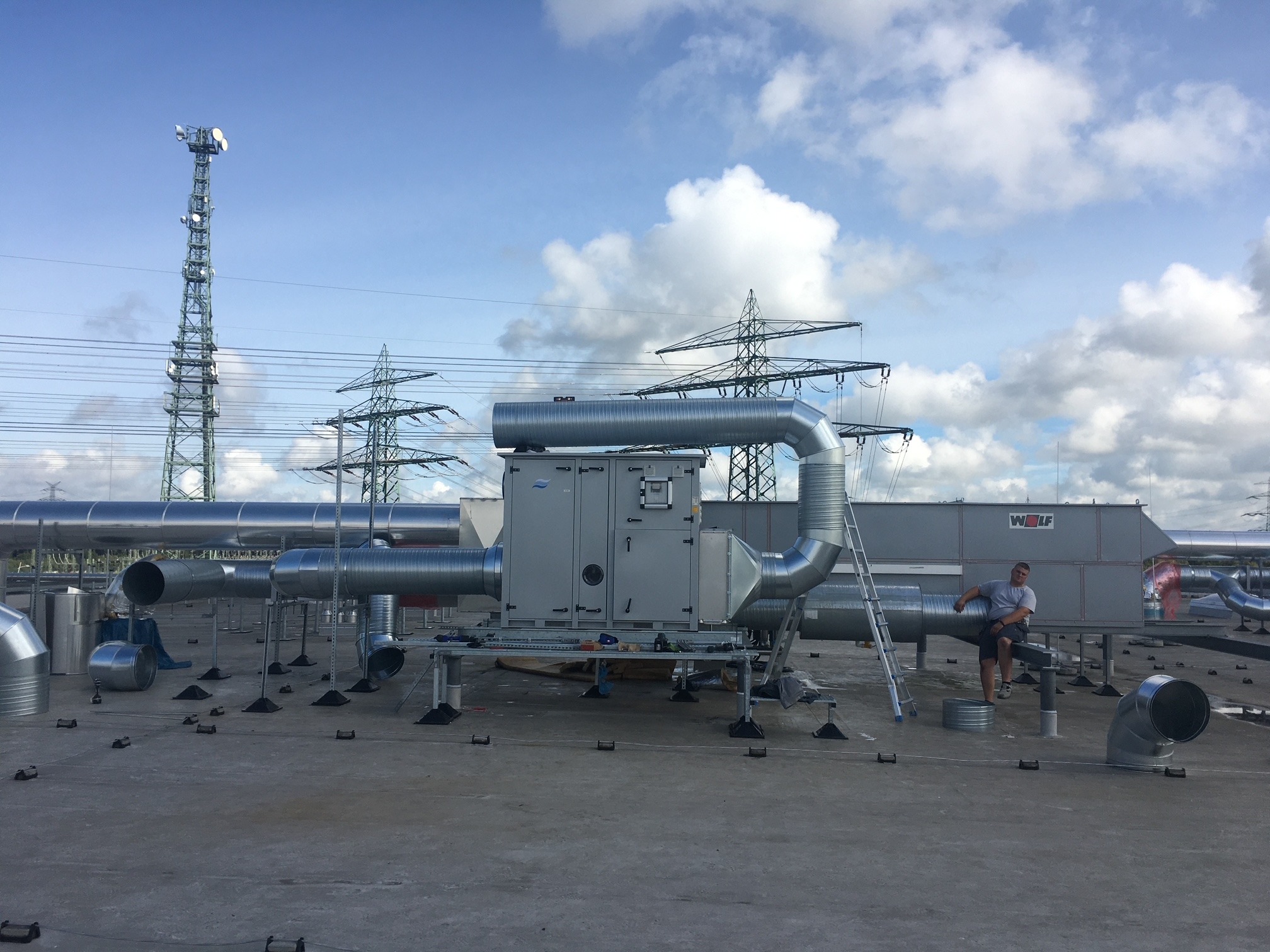 Instalacja wentylacyjna na dachu budynku z widoczną wieżą telekomunikacyjną i liniami wysokiego napięcia w tle. Mężczyzna siedzi na rurze wentylacyjnej.