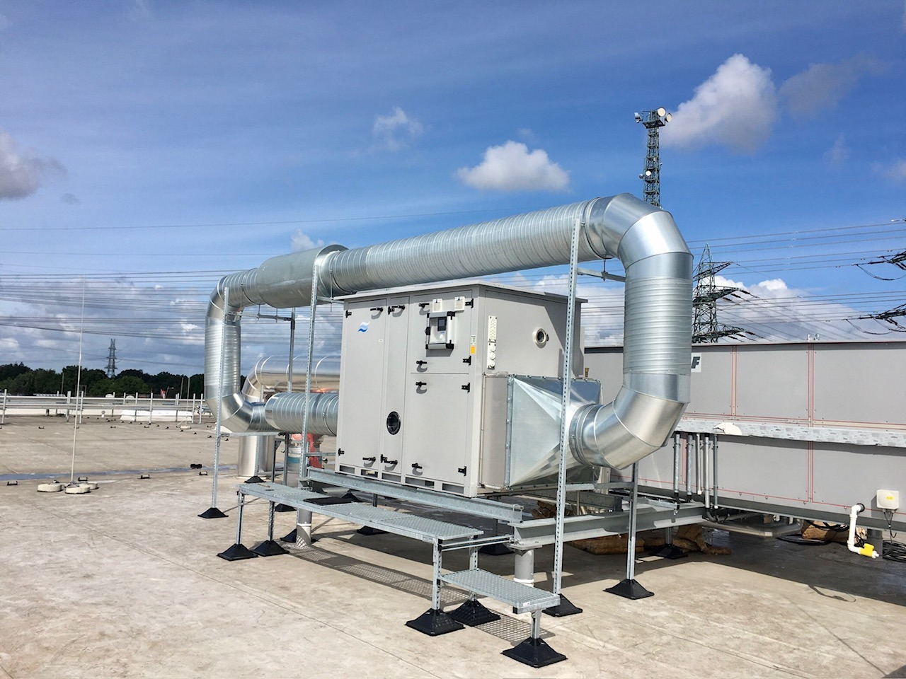 Instalacja wentylacyjna na dachu budynku, z widocznymi przewodami wentylacyjnymi, wspornikami i elementami mocującymi. W tle błękitne niebo z chmurami.