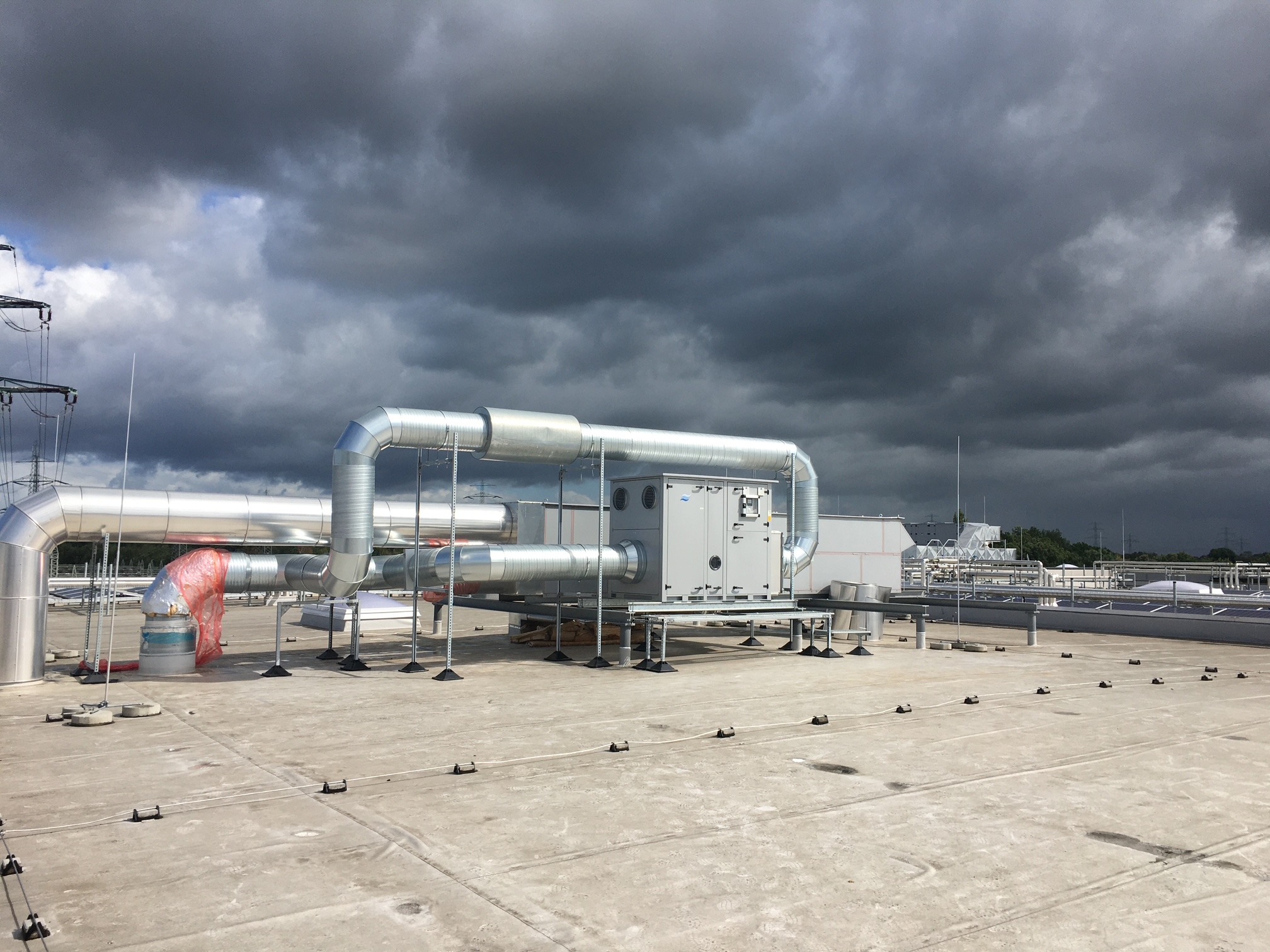 Instalacja przemysłowa z srebrnymi rurami i szarą centralą wentylacyjną na płaskim dachu, widok z góry, pochmurne niebo w tle.