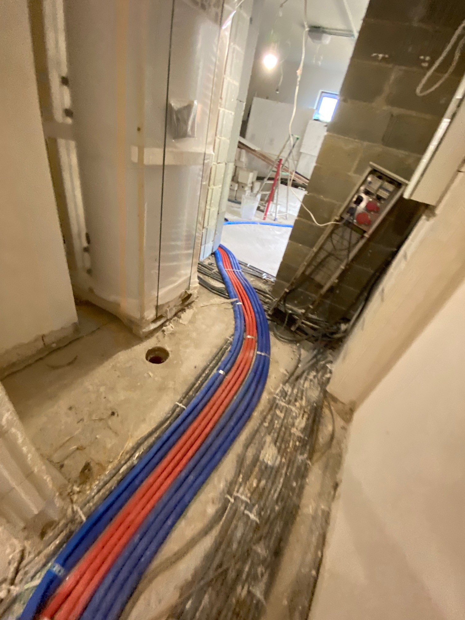 Wnętrze w trakcie instalacji: rury grzewcze czerwone i niebieskie ułożone wzdłuż ściany, widoczne także inne instalacje i elementy konstrukcyjne.