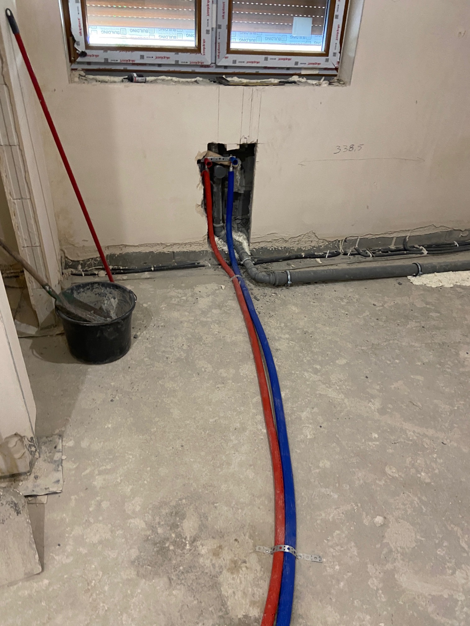 Instalacja wodna w trakcie montażu: czerwona i niebieska rura prowadzone w ścianie. Widoczne narzędzia i elementy mocujące. Surowy stan wykończenia.