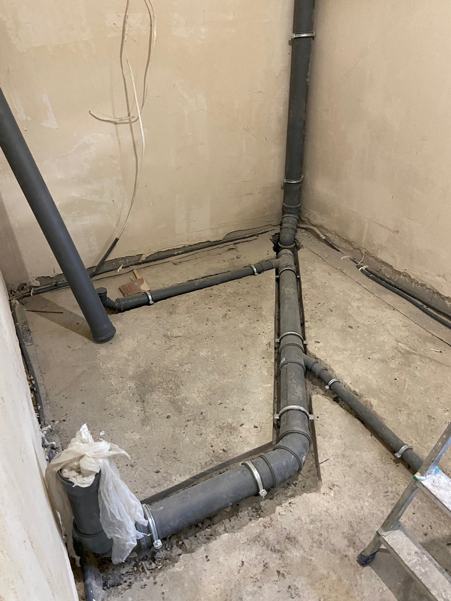 Szare rury kanalizacyjne z obejmami, ułożone w pomieszczeniu w trakcie prac instalacyjnych. Część rur zagłębiona w podłodze, widoczny fragment drabiny.