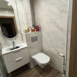 Krzysztof Stachnio - Jasna łazienka po renowacji: białe marmurowe płytki, drewnopodobna podłoga, owalne lustro, biała szafka z umywalką, toaleta i akcesoria.