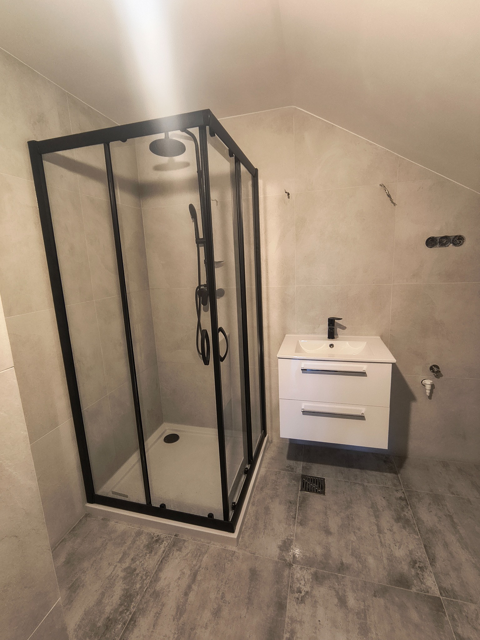 Nowoczesna łazienka w odcieniach szarości z kabiną prysznicową typu walk-in z czarnymi profilami i białą szafką pod umywalką z dwiema szufladami.